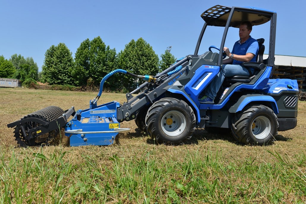 man driving a mini loader with a power harrow on a field