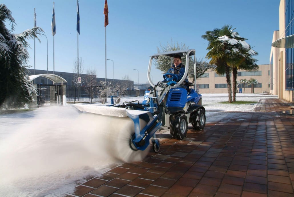 a mini loader with rotary broom cleaning a snowy pavement