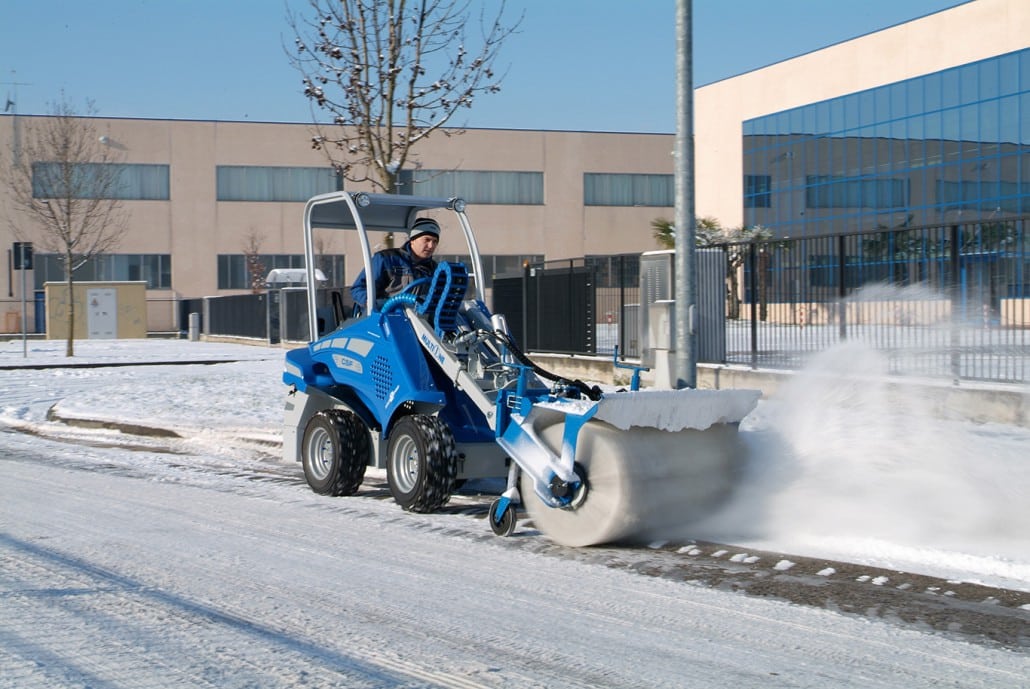 a man driving a mini loader with a rotary broom attached, cleaning a snowy street